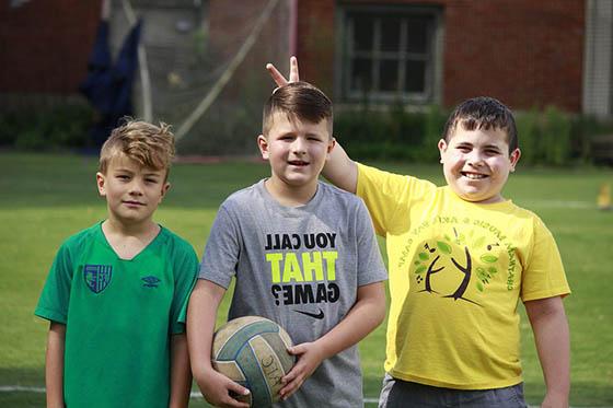 三个男孩拿着足球在球场上摆姿势，对着镜头微笑. 一个男孩竖起手指，对另一个男孩竖起耳朵.